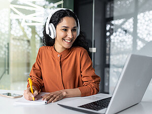 Junge Frau mit Kopfhörer am Laptop Latin american business woman with curly hair and headphones watching online training course at workplace, woman writing information happy and satisfied with the results of professional development